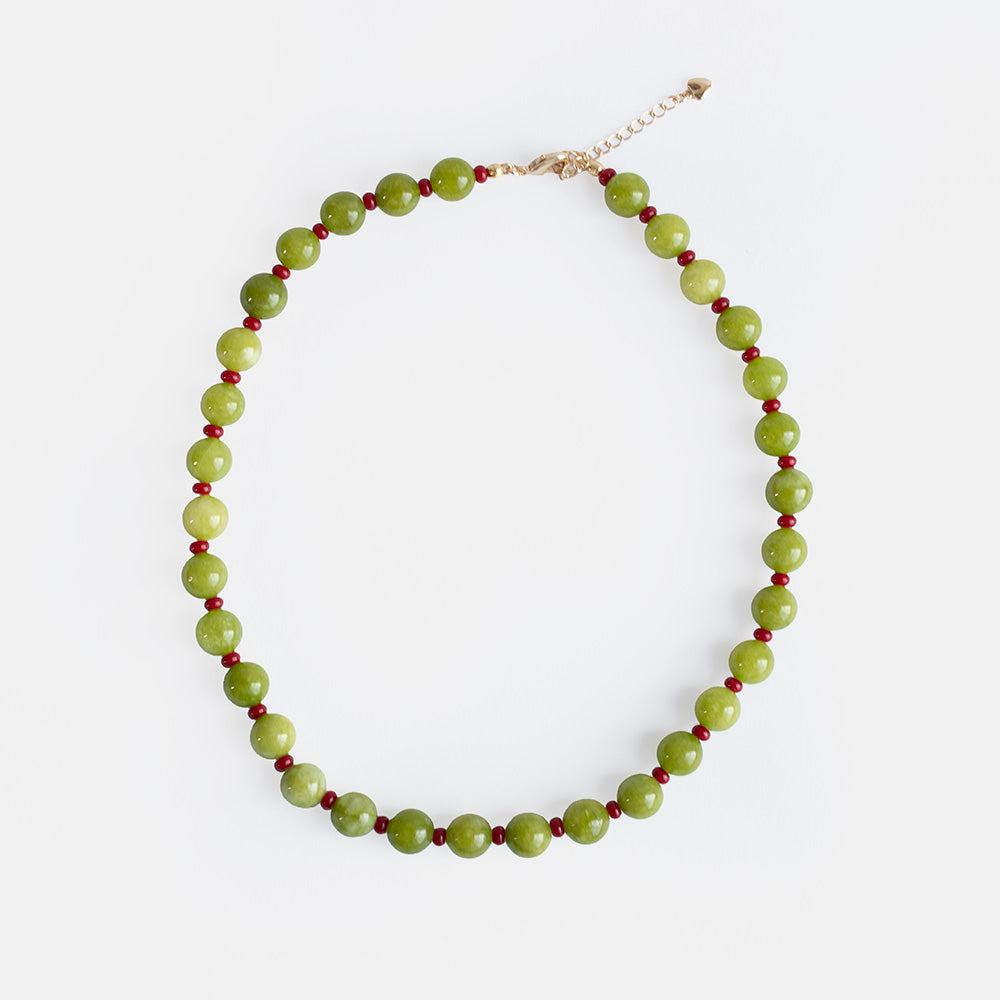 A handmade beaded necklace of round olive green semi-precious stone beads alternating with small red beads, with a gold-tone clasp and extender chain, on a light grey background.