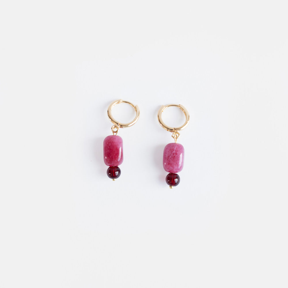 A pair of small gold huggie hoop earrings each with a raspberry pink semi-precious stone bead above a small burgundy bead, on a light grey background.