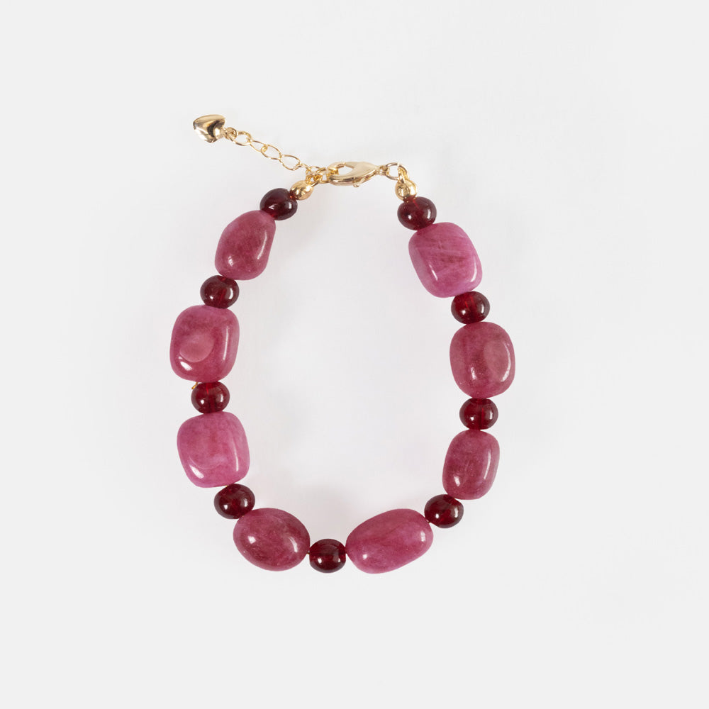 A handmade bracelet of chunky raspberry pink semi-precious stone beads alternating with round burgundy beads, with a gold-tone clasp, extender chain and heart charm, on a light grey background.