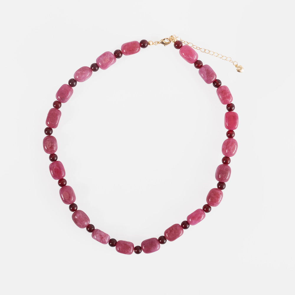 A beaded necklace featuring faceted pinkish-red beads separated by small dark red beads, with a gold-tone clasp and extender chain, displayed on a light gray background.