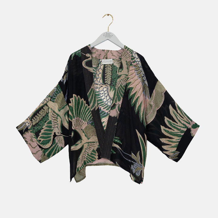 A black open-front kimono-style top with wide three-quarter sleeves, featuring a detailed botanical and bird pattern in green, beige, and dusty pink. The garment is displayed on a white hanger against a light gray background.