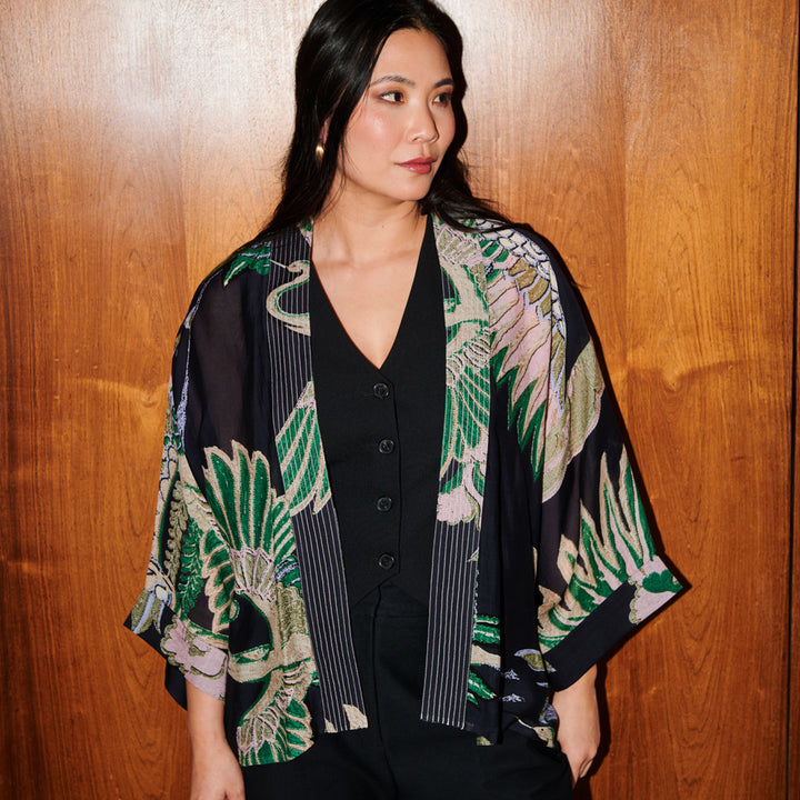 A woman wearing a black open-front kimono with a green, pink, and beige tropical bird and leaf print, layered over a black button-up top, standing against a wooden wall.