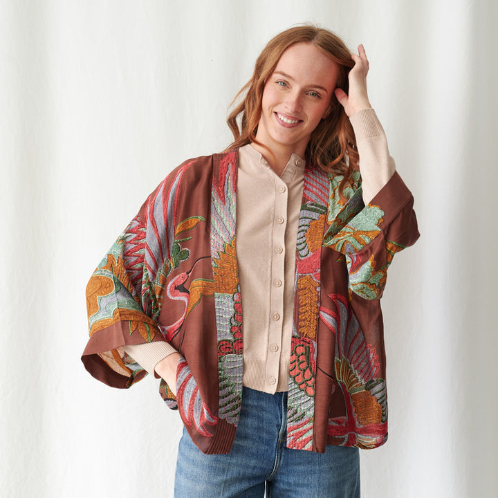 A woman wearing a brown open-front short kimono with a green, pink, and blue tropical bird and leaf print, layered over a beige button-up top, standing against a light grey wall.