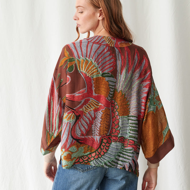 A woman wearing a brown short kimono with a green, pink, and blue tropical bird and leaf print, worn with light denim jeans against a white background.
