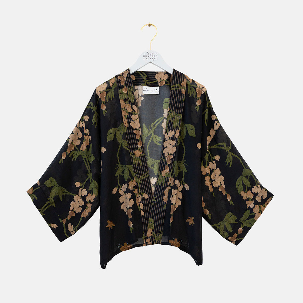 A black open-front kimono-style cardigan with wide three-quarter sleeves, featuring a green and beige floral print and vertical pleats down the front, hanging on a white hanger against a light gray background.