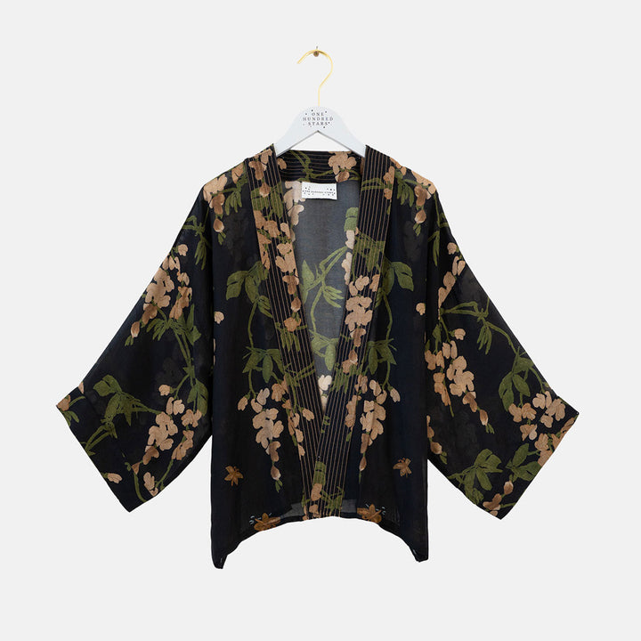 A black open-front kimono-style cardigan with wide three-quarter sleeves, featuring a green and beige floral print and vertical pleats down the front, hanging on a white hanger against a light gray background.