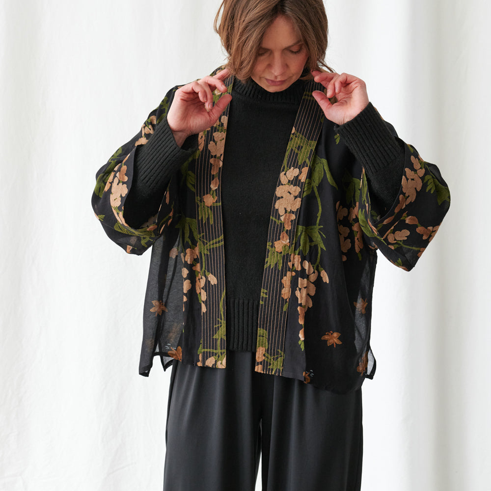A woman wearing a black open-front cardigan with a beige and green floral print, layered over a black ribbed turtleneck sweater and black pants, posing against a white curtain background.