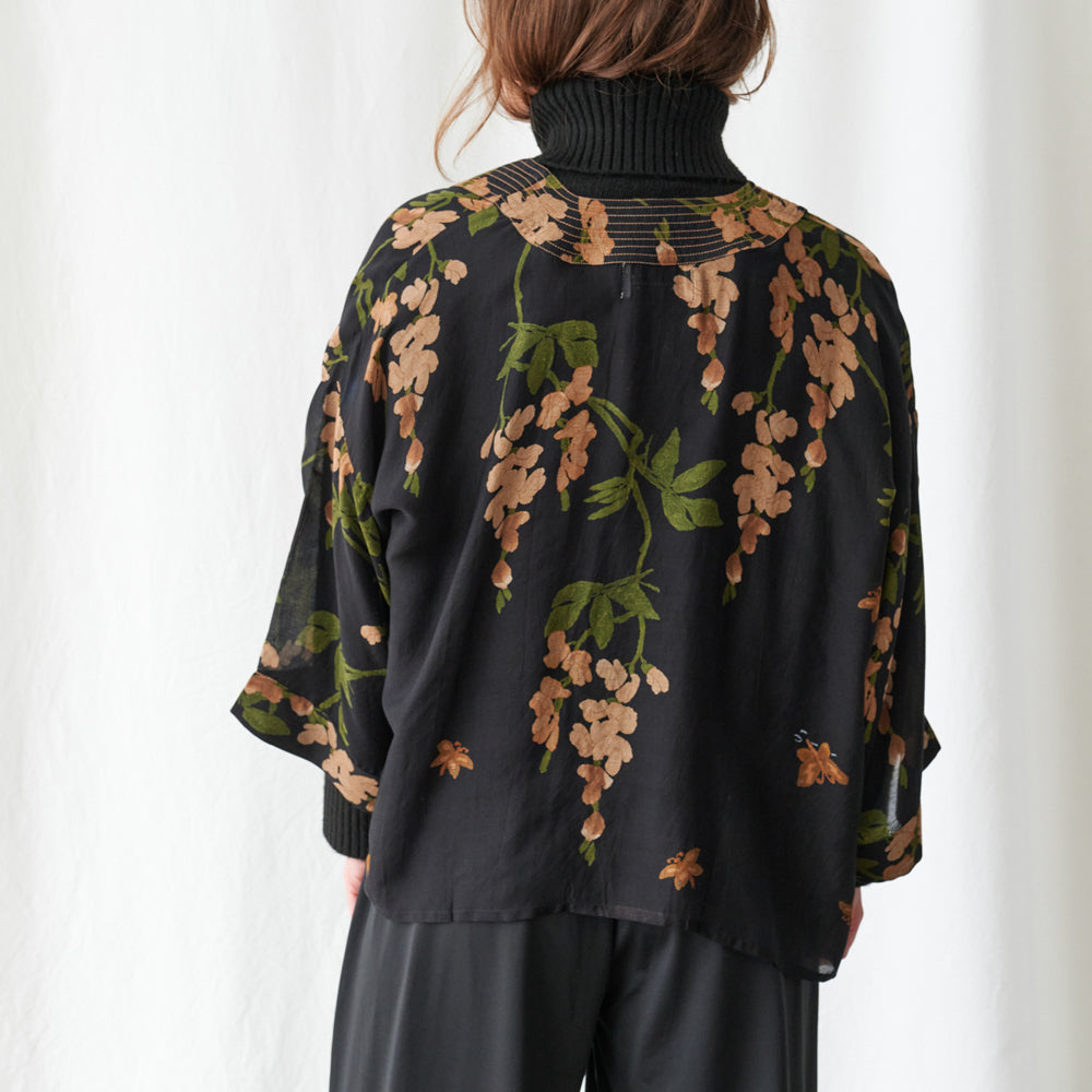 A woman viewed from behind wearing a black turtleneck top with a sheer mesh back panel and a black overlay jacket featuring a brown and green floral wisteria print, paired with black pants against a white curtain background.