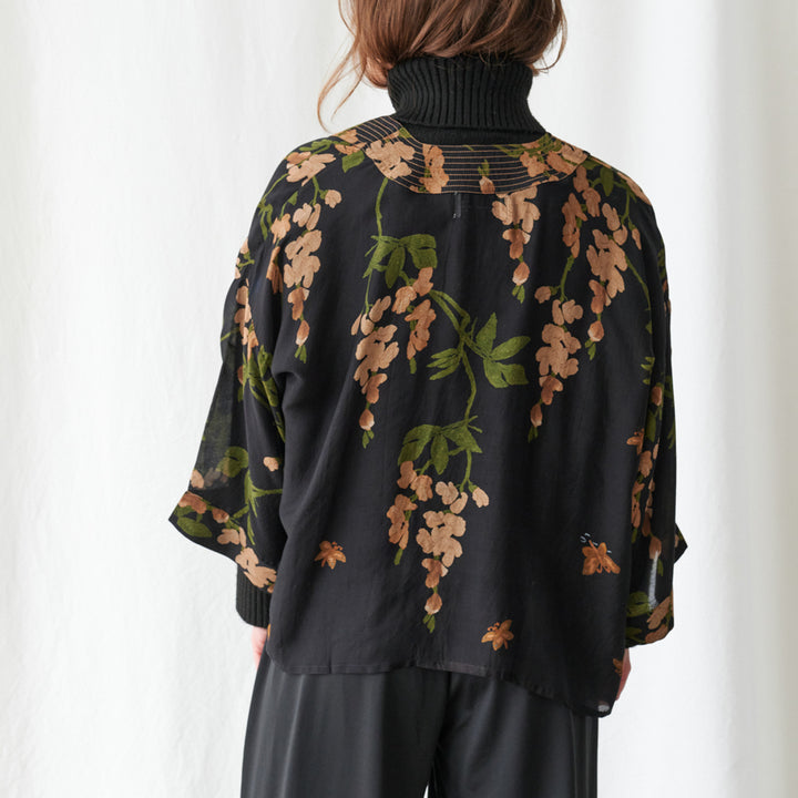 A woman viewed from behind wearing a black turtleneck top with a sheer mesh back panel and a black overlay jacket featuring a brown and green floral wisteria print, paired with black pants against a white curtain background.