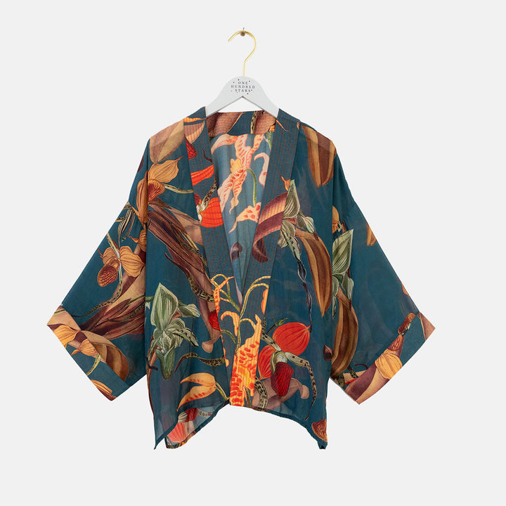 A lightweight open-front kimono-style top with wide sleeves, featuring a vibrant multicolor print of orchid plants on a dark teal-blue background. The garment is displayed on a white hanger with a gold hook against a solid light gray background.