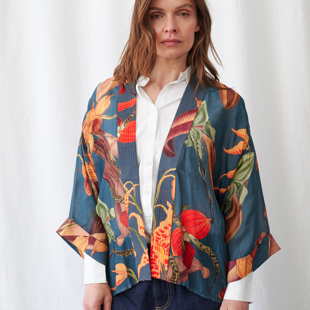 A woman wearing a teal short wool kimono with a bold orange and plum orchid print, layered over a white shirt with dark jeans, against a white curtain.