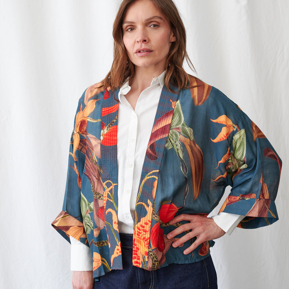 A woman wearing a blue open-front kimono-style jacket with a vibrant tropical floral print, layered over a white button-down shirt and dark blue jeans, standing against a white curtain backdrop.
