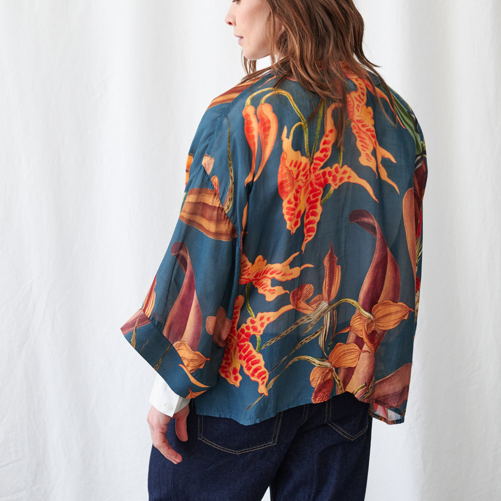 A woman shown from the back wearing a loose, short-sleeved blouse with a bold floral print in orange, red, and yellow on a dark teal-blue background, paired with dark blue jeans against a white fabric backdrop.