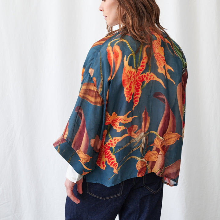 A woman shown from the back wearing a loose, short-sleeved blouse with a bold floral print in orange, red, and yellow on a dark teal-blue background, paired with dark blue jeans against a white fabric backdrop.