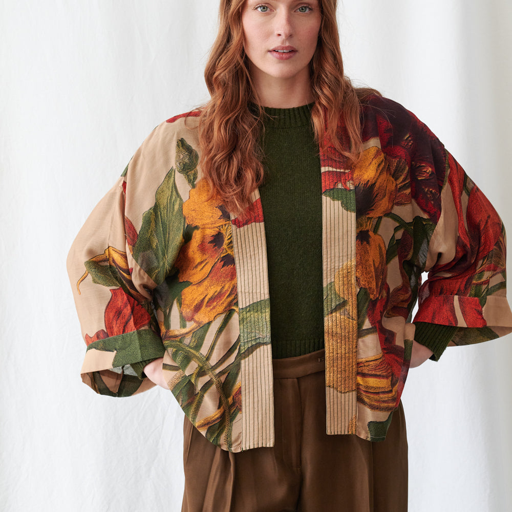 A woman wearing a beige open-front jacket with a bold red, orange, and green floral print, layered over a dark green knit sweater and brown pants, photographed against a white studio background.