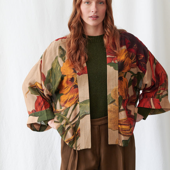 A woman wearing a beige open-front jacket with a bold red, orange, and green floral print, layered over a dark green knit sweater and brown pants, photographed against a white studio background.