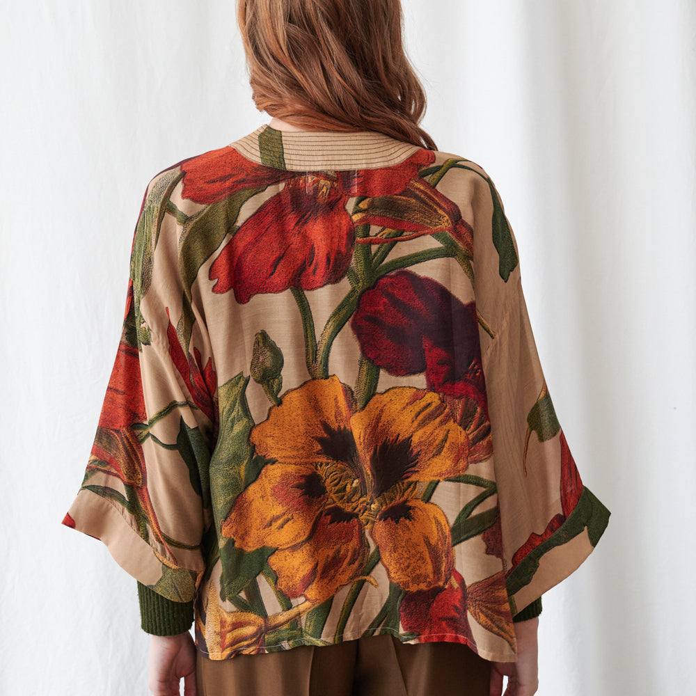 A woman viewed from behind wearing a beige kimono-style cardigan with a large floral print featuring red, orange, and yellow flowers with green leaves, set against a plain white background.