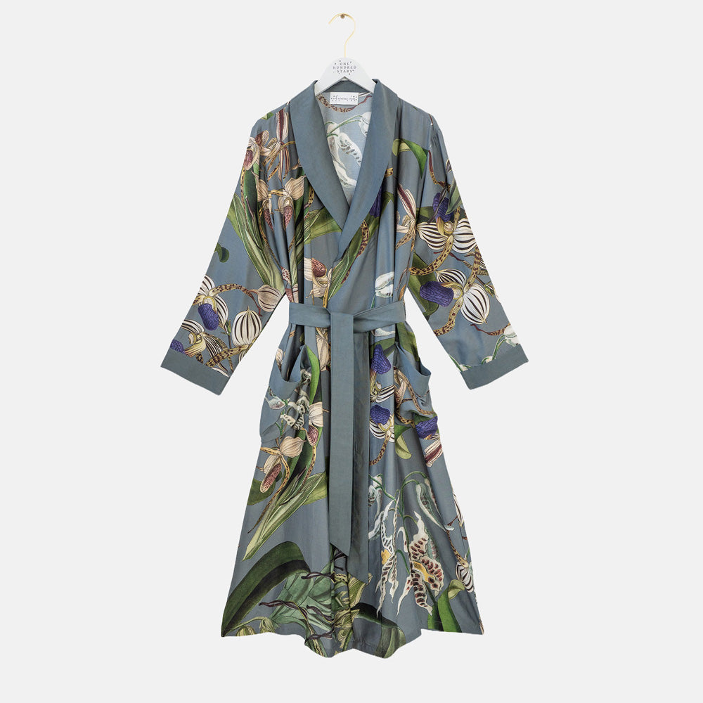 A long-sleeved wrap robe with a wide collar and self-tie belt, featuring an all-over botanical print on a muted blue-gray base. The print includes green leaves, purple flowers, and striped patterns.