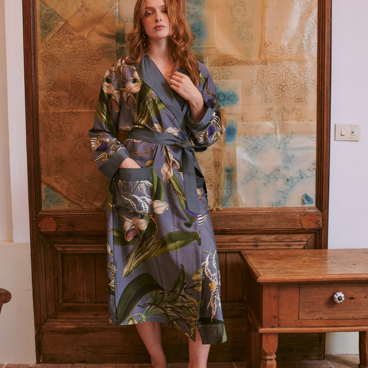 woman wearing a grey orchid print dressing gown with purple, green and white detailing stood in front of a wooden dresser and table