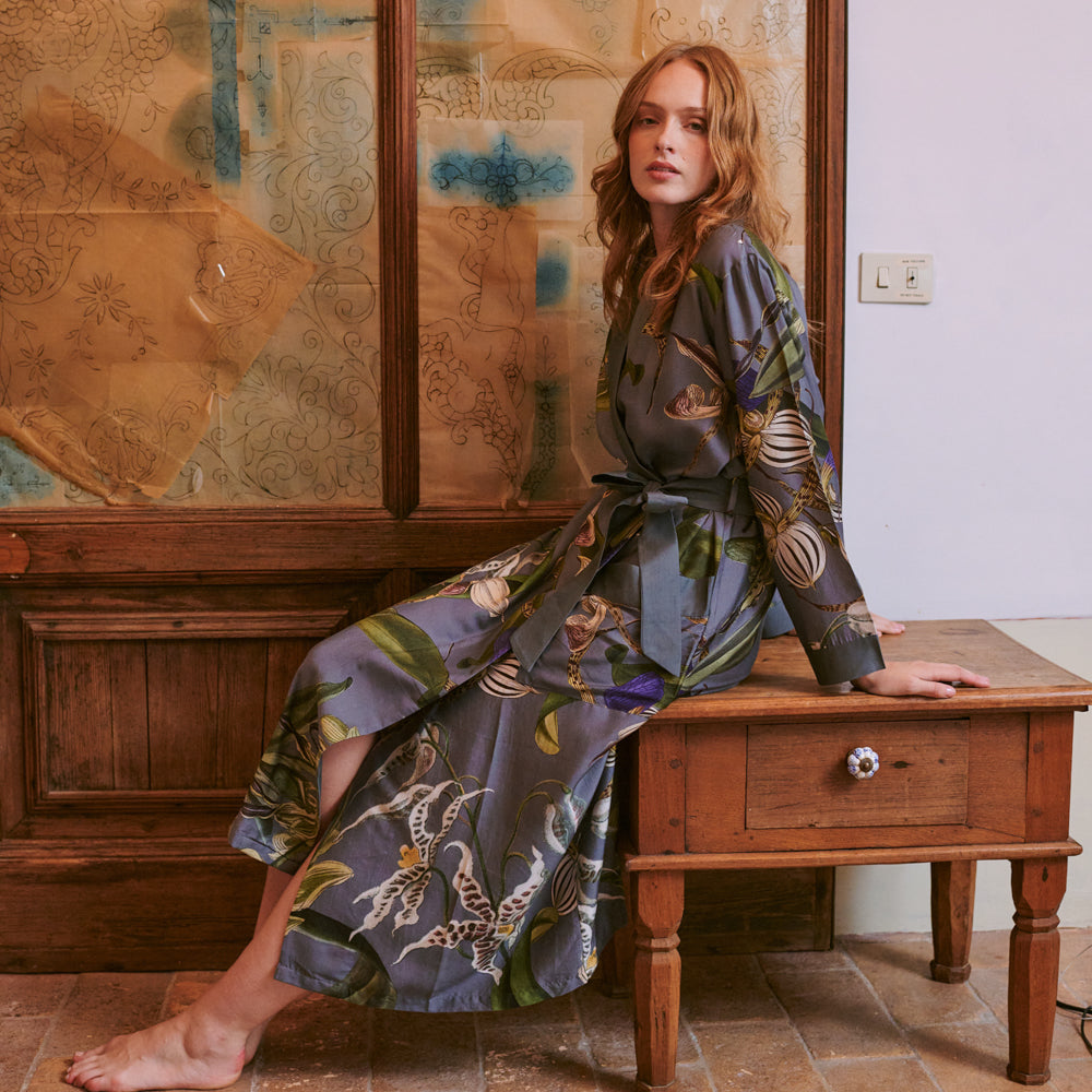 woman wearing a grey orchid print dressing gown with purple, green and white detailing sat on a wooden table