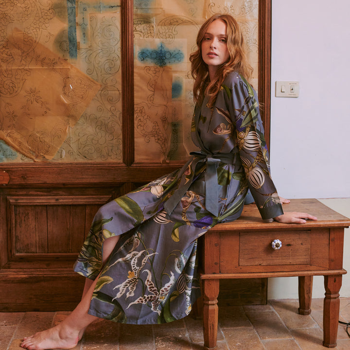 woman wearing a grey orchid print dressing gown with purple, green and white detailing sat on a wooden table