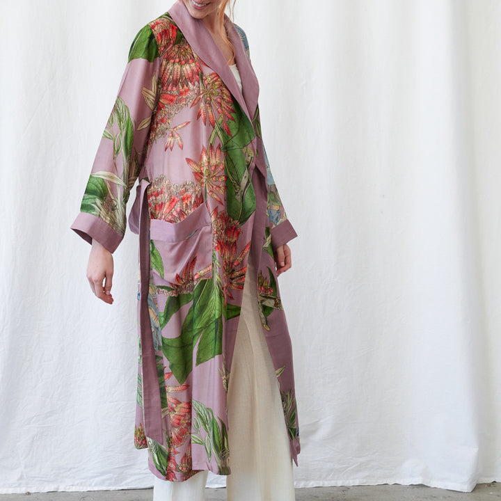 A woman models a long, open-front robe with a dusty purple base and a tropical floral print featuring green leaves and red-orange flowers, styled over a cream dress against a white fabric backdrop.