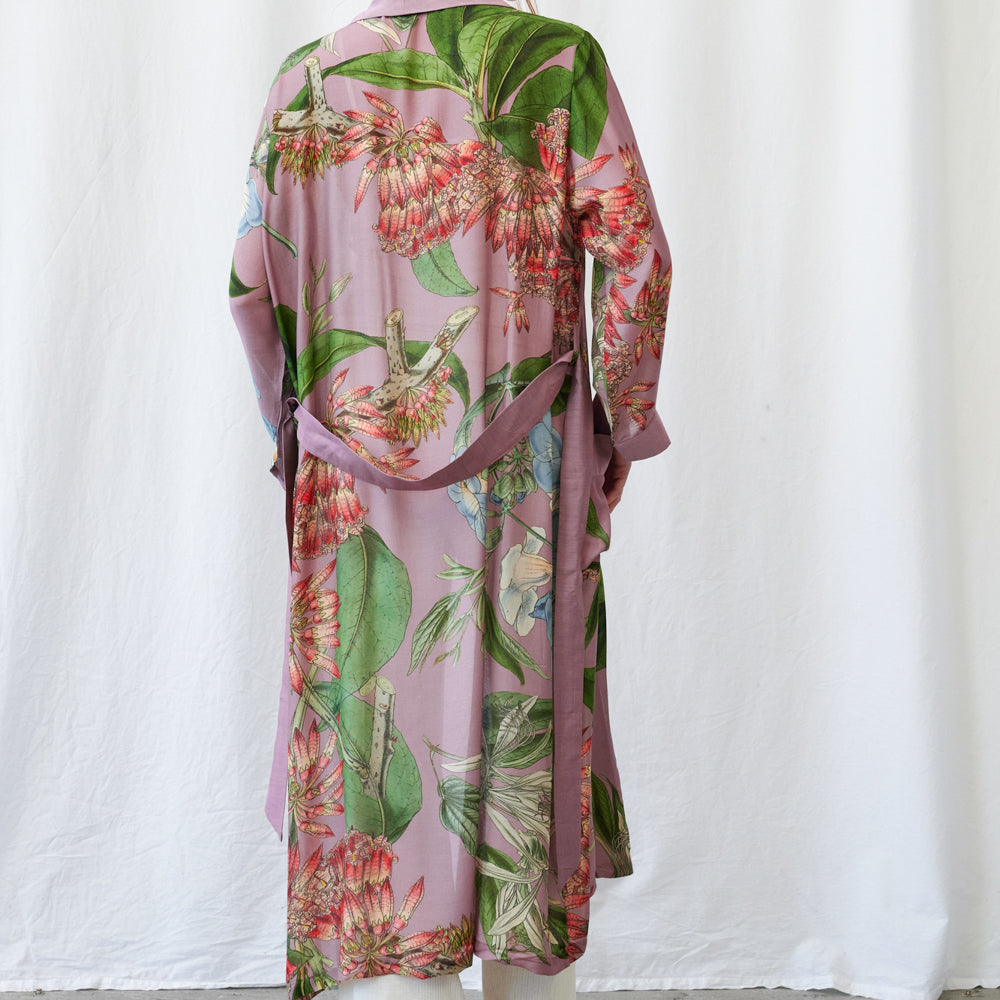 A long-sleeved wrap robe with a pink base and a tropical floral print, shown from the back on a mannequin against a white fabric backdrop.
