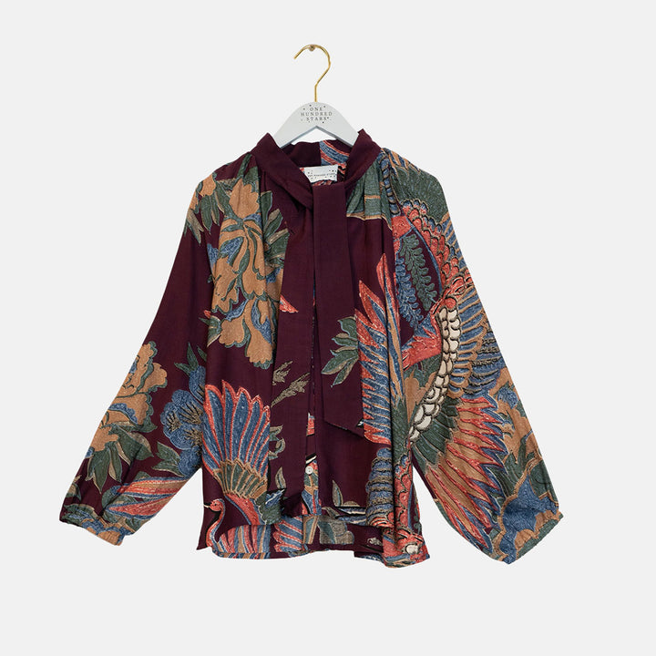 A long-sleeve blouse with a split design, featuring a deep burgundy front panel with a tie-neck bow and a contrasting multicolor batik patterned back and sleeves. The blouse is displayed on a white hanger against a light gray background.