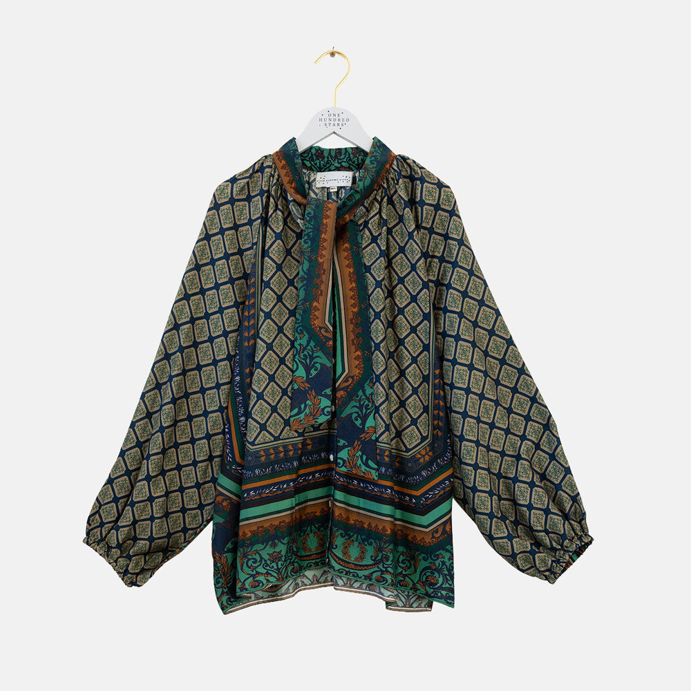 A long-sleeve blouse with a geometric and paisley pattern in green, blue, and brown, hanging on a white hanger against a light gray background.