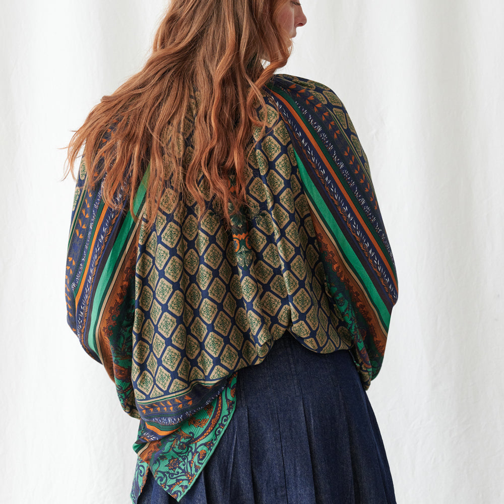 A woman shown from the back wearing a patterned shawl draped over her shoulders and tied at the waist, paired with a dark blue skirt. The shawl features a complex geometric and tribal-inspired pattern in navy blue, olive green, rust orange, and cream.