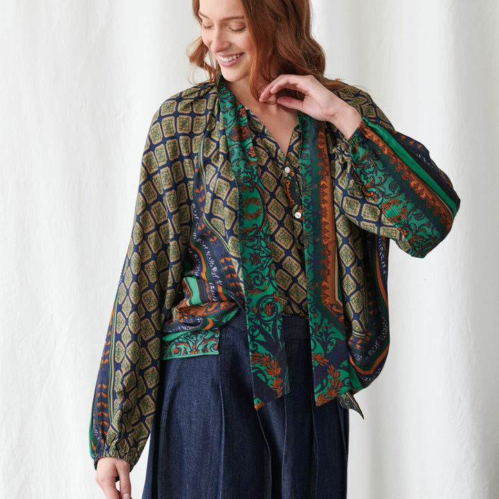 A woman wearing a patterned shirt draped  paired with a dark blue skirt. The blouse features a complex geometric and tribal-inspired pattern in navy blue, olive green, rust orange, and cream.