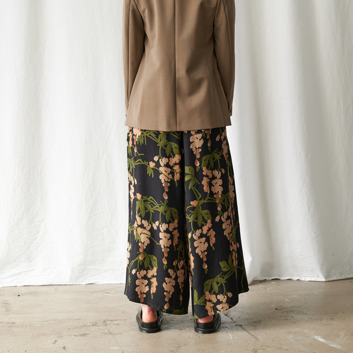 A person shown from the back wearing a tan blazer and wide-leg black skirt with a beige floral print, standing against a white draped fabric backdrop.