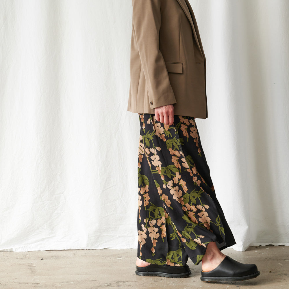 A person shown from the waist down wearing a tan blazer, a black wide-leg skirt with a peach and green floral print, and black clogs, standing against a white draped fabric backdrop.