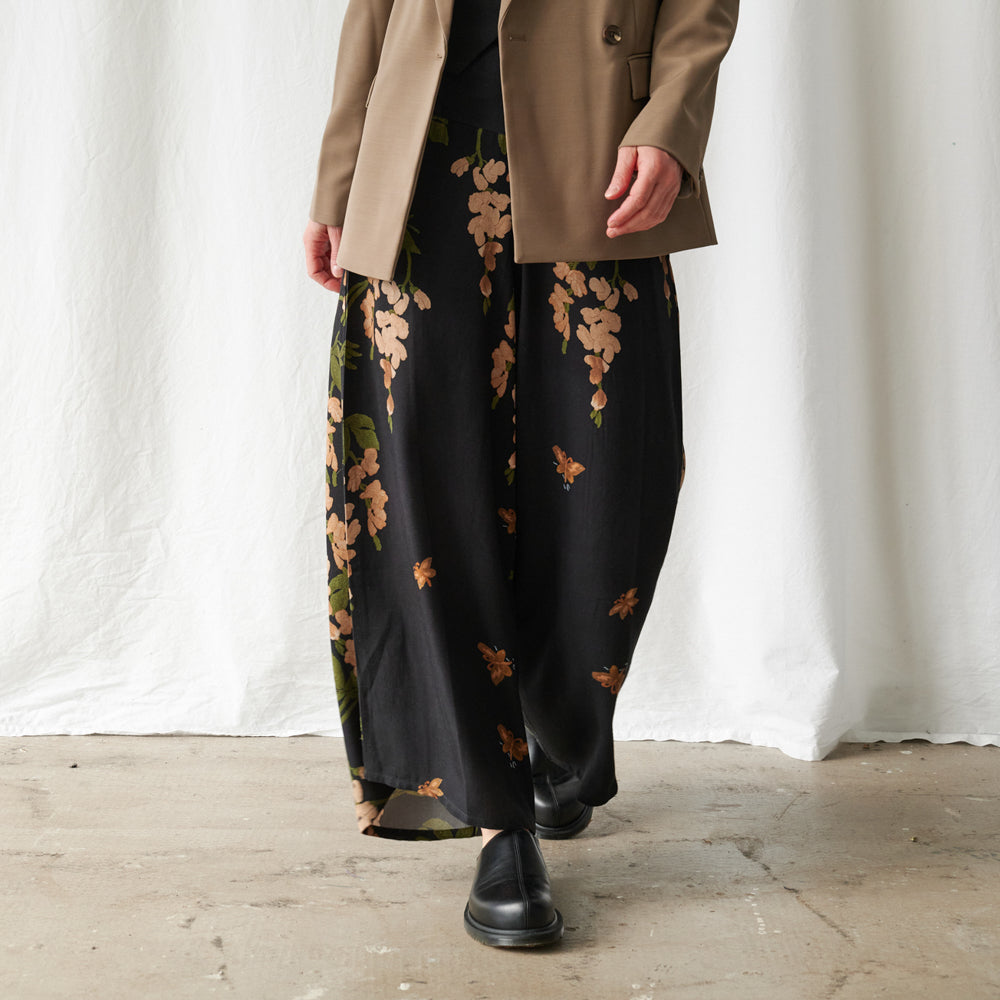 A person wearing wide-leg black pants with a tan and green floral print, styled with a tan blazer and black boots, standing against a white draped fabric backdrop.
