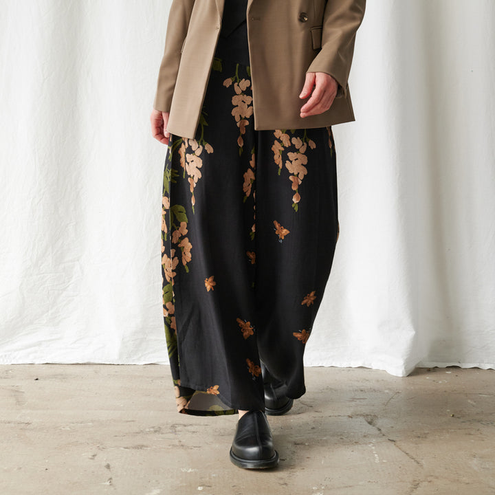 A person wearing wide-leg black pants with a tan and green floral print, styled with a tan blazer and black boots, standing against a white draped fabric backdrop.