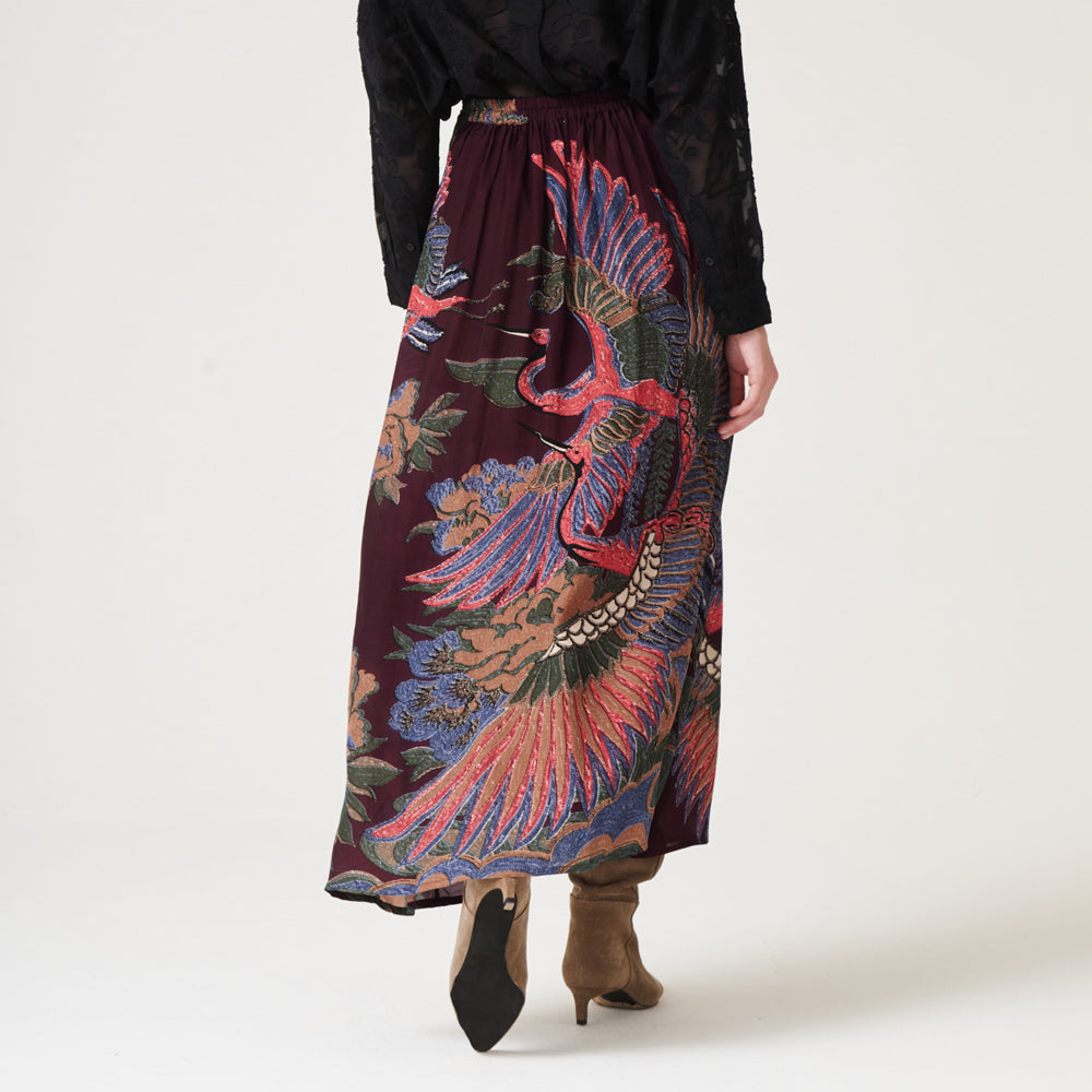 woman wearing an a-line maxi skirt featuring a colourful pink, green & blue cranes design on a dark purple base with a black shirt tucked in and brown suede boots