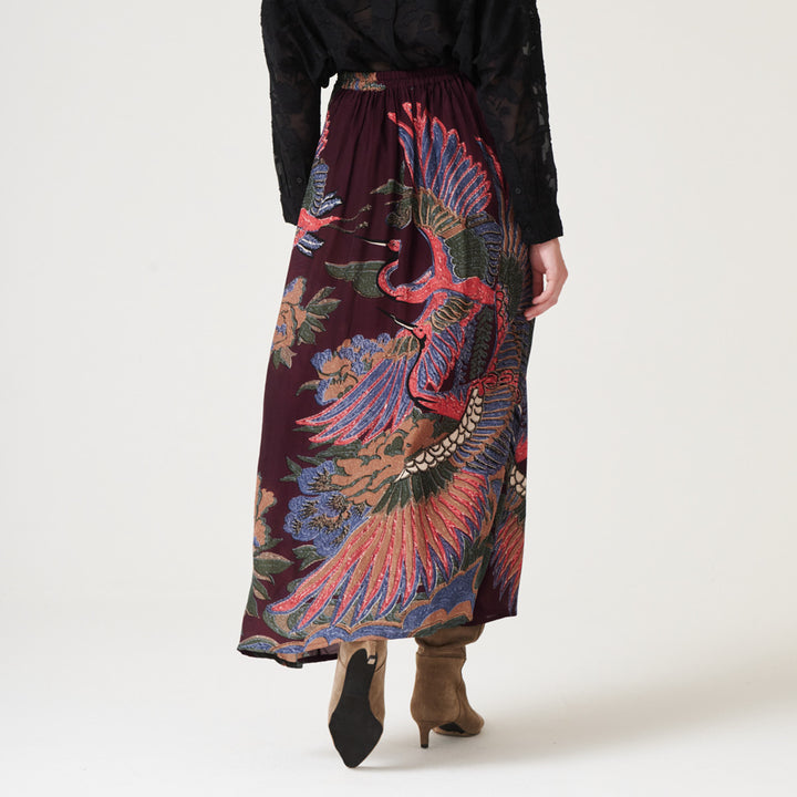 woman wearing an a-line maxi skirt featuring a colourful pink, green & blue cranes design on a dark purple base with a black shirt tucked in and brown suede boots