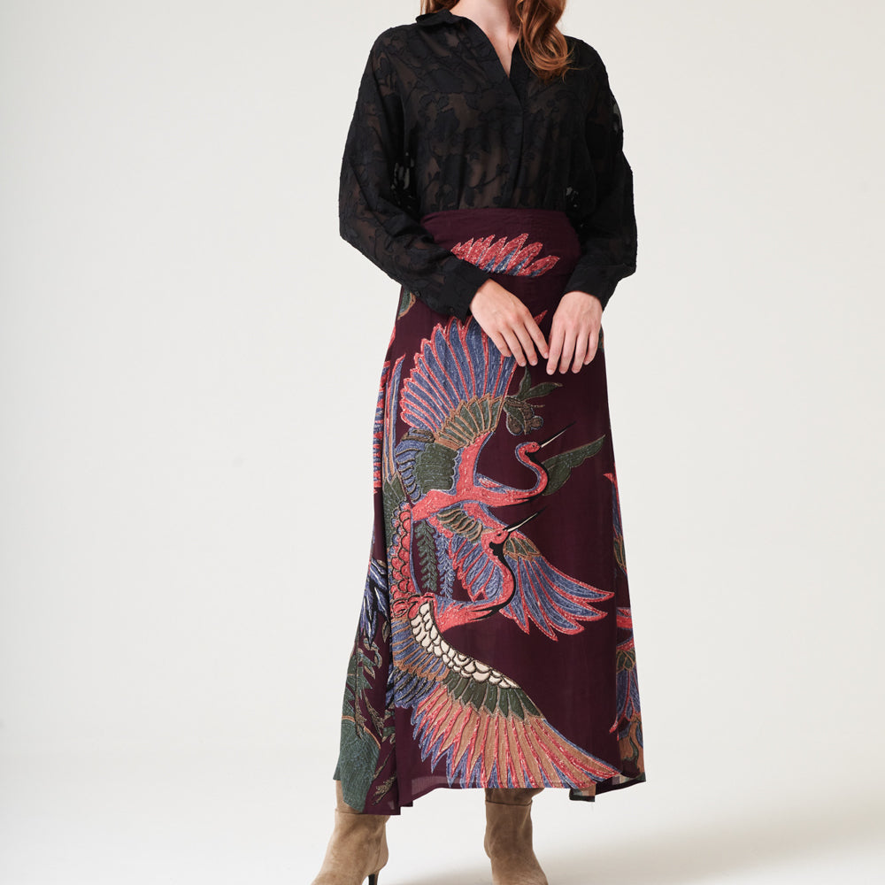 woman wearing an a-line maxi skirt featuring a colourful pink, green & blue cranes design on a dark purple base with a black shirt tucked in and brown suede boots