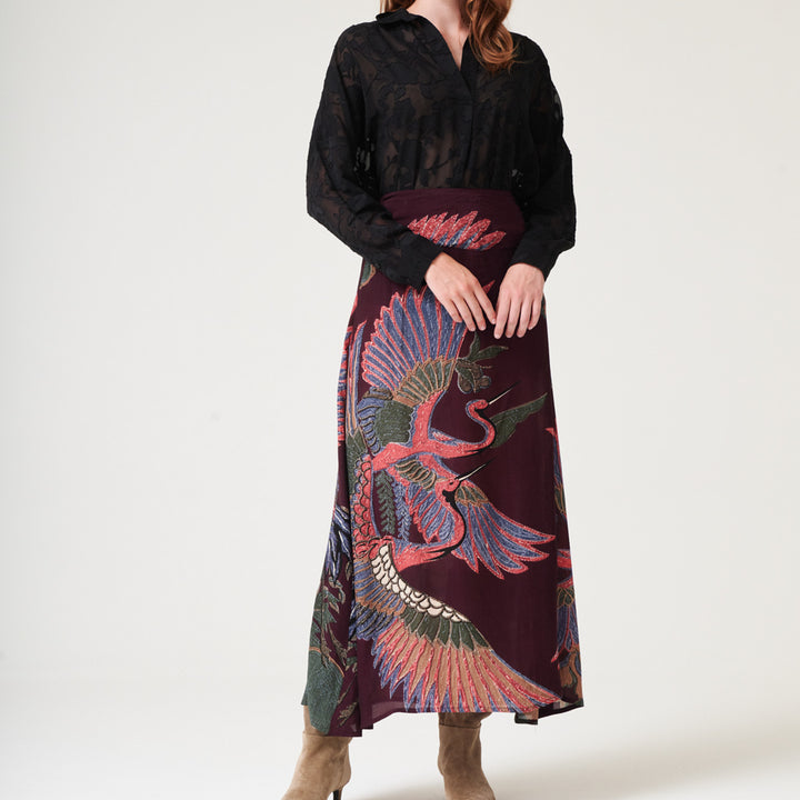 woman wearing an a-line maxi skirt featuring a colourful pink, green & blue cranes design on a dark purple base with a black shirt tucked in and brown suede boots