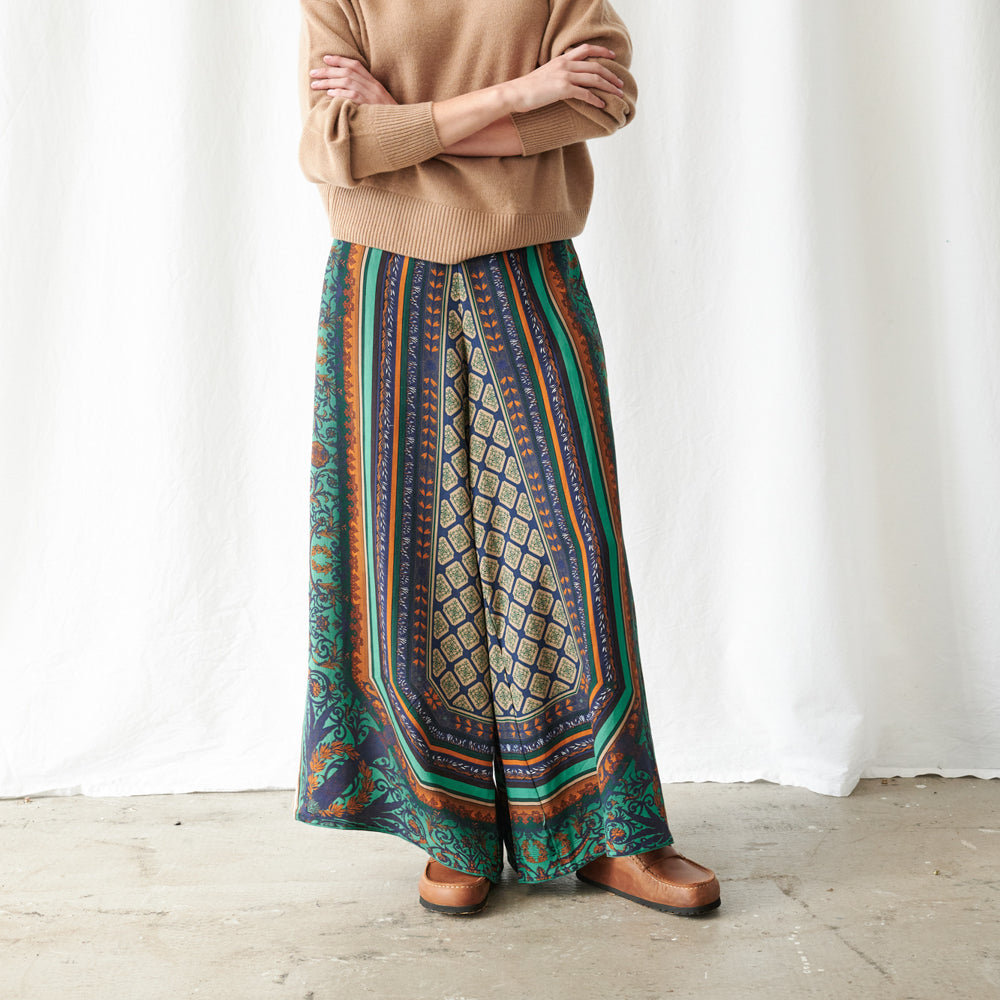 A person wearing a long, wide-leg maxi skirt with a complex geometric and tribal-inspired pattern in teal, navy, rust orange, and beige, styled with a tan sweater and brown shoes against a white curtain backdrop.