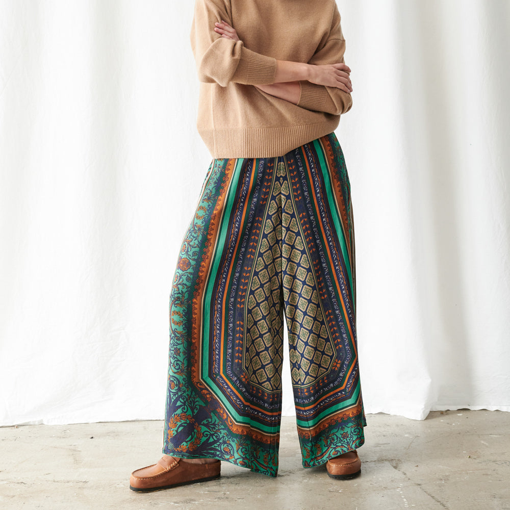 A person wearing wide-leg patterned pants with a beige sweater and brown shoes, standing against a white curtain backdrop.