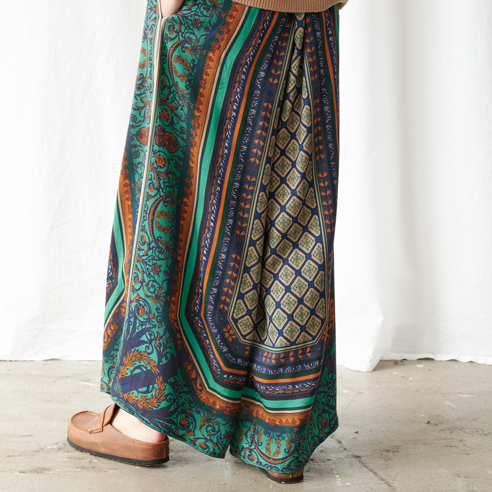 A person wearing a long, wide-leg skirt with a complex multicolor pattern featuring teal, navy, orange, and gold, paired with brown leather loafers, standing against a white curtain backdrop.
