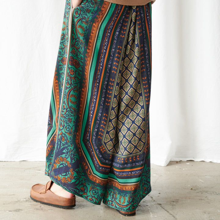 A person wearing a long, wide-leg skirt with a complex multicolor pattern featuring teal, navy, orange, and gold, paired with brown leather loafers, standing against a white curtain backdrop.