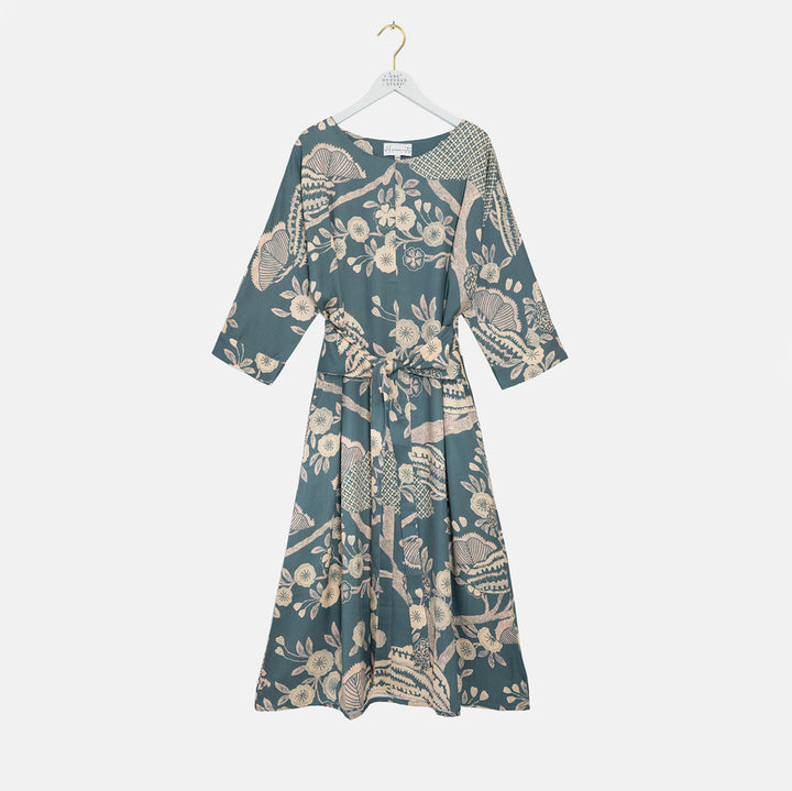 A three-quarter sleeve midi dress with a tie waist, featuring an all-over botanical pattern in beige on a muted teal background, hanging on a white hanger against a light gray background.