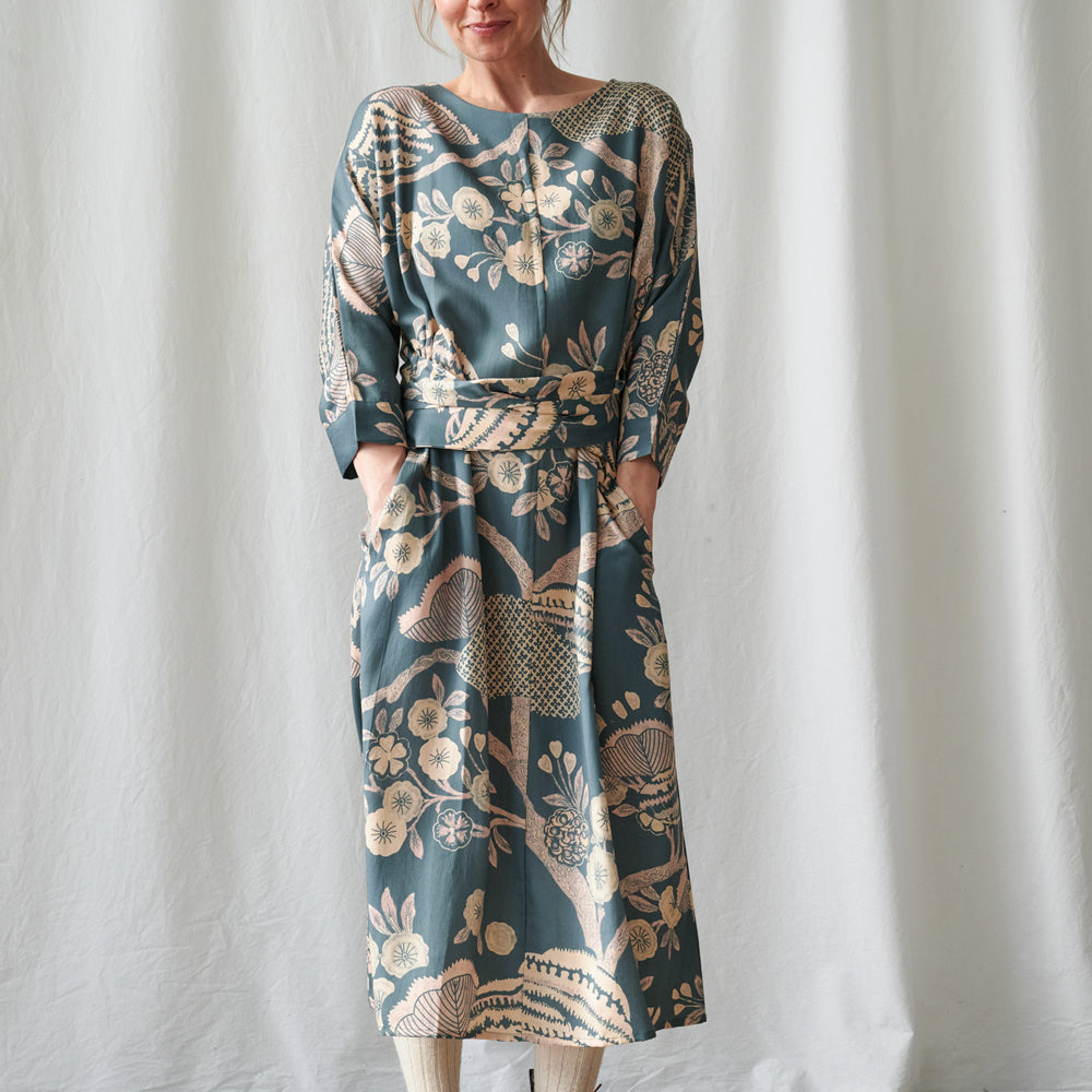 A woman wearing a long-sleeved midi dress with a botanical pattern in muted teal, beige, and brown, standing against a white curtain backdrop.