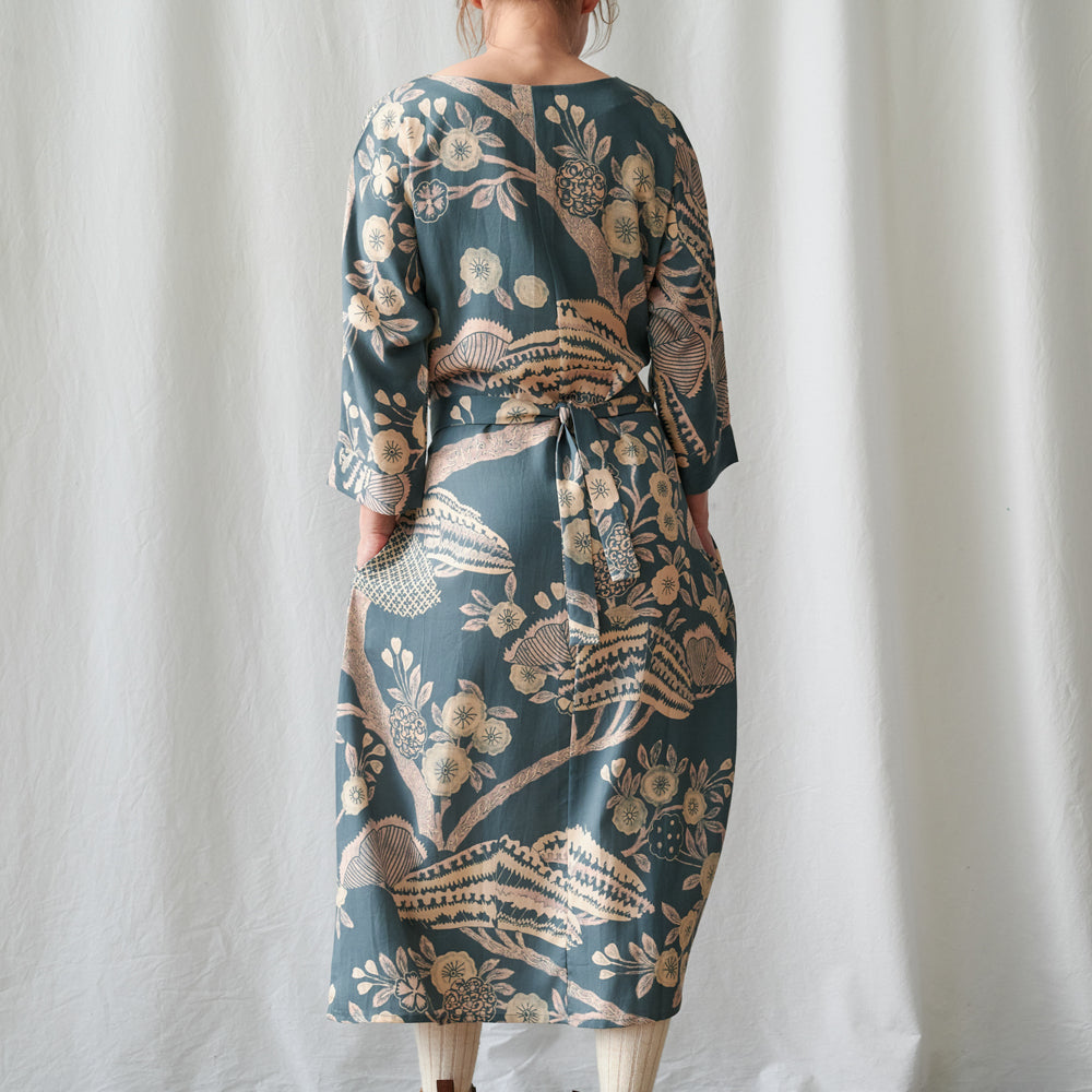 A woman wearing a long-sleeved midi dress with a botanical pattern, shown from the back against a white curtain backdrop.