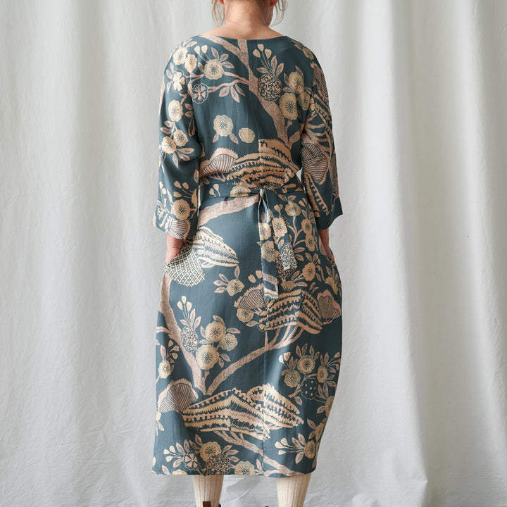 A woman wearing a long-sleeved midi dress with a botanical pattern, shown from the back against a white curtain backdrop.