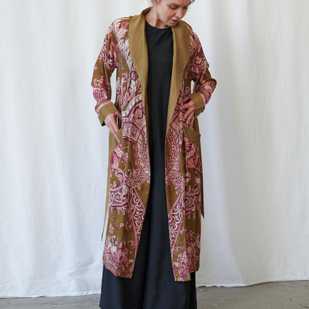 A woman wearing a long open-front kimono-style gown with a pink and brown paisley pattern and a solid tan collar, worn over a black dress, standing against a white fabric backdrop.
