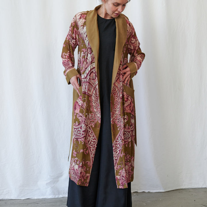 A woman wearing a long open-front kimono-style gown with a pink and brown paisley pattern and a solid tan collar, worn over a black dress, standing against a white fabric backdrop.