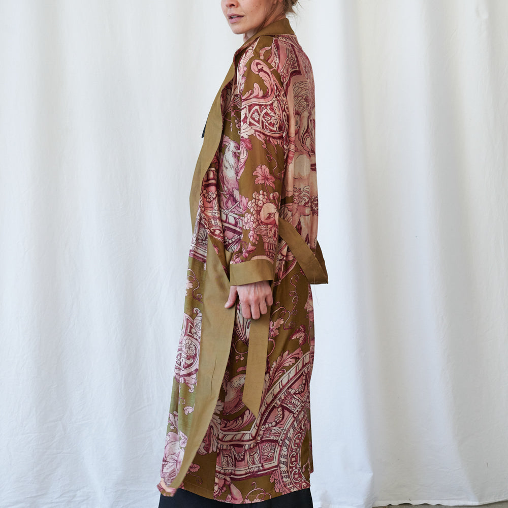 A model shown from the side wearing a long-sleeved, open-front kimono-style robe with a brown base and pink paisley pattern, set against a white draped fabric backdrop.